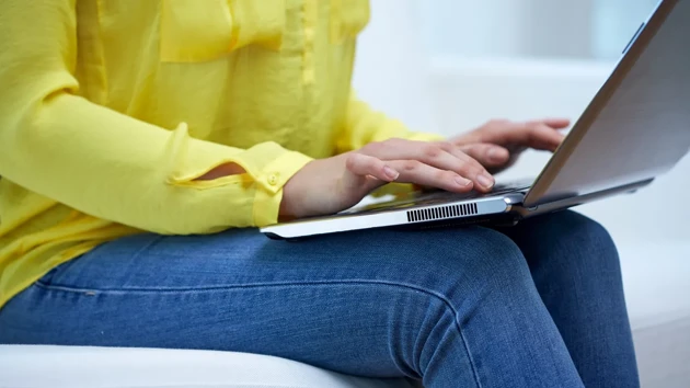 Detaljbild på kvinna i soffa som sitter med uppslagen laptop i en soffa och skriver.