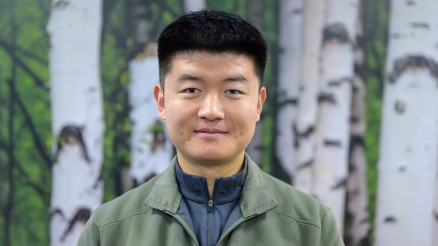 A man with a green zip up sweater poses in front of a birch tree background
