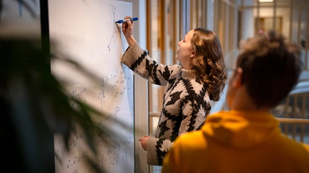 A woman is writing formulas on a whiteboard while a man is watching.