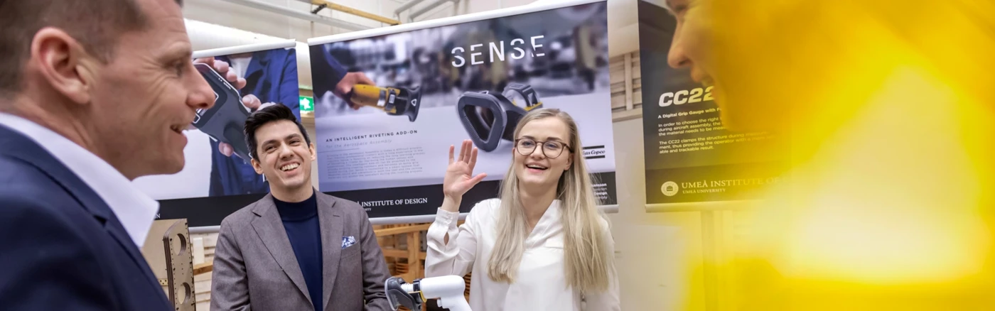 en glad ung kvinna samtalar med tre män, företagsrepresentanter. De står runt ett podium med en modell på. i bakgrunden syns stora affischer med bildmaterial på.