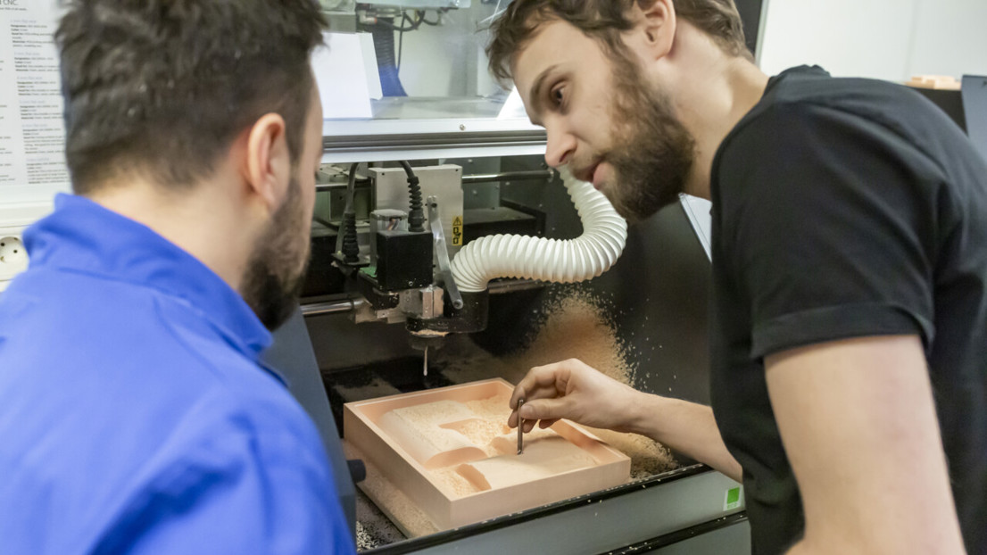 Två personer står framåtlutade över en verkstadsmaskin (3D-fräs). Vi ser dem bakifrån i närbild. Den ene tittar på den andre och håller ett frässtål i högra handen. Han liksom pekar med frässtålet och man förstår av situationen att han förklarar någo för den andra personen.